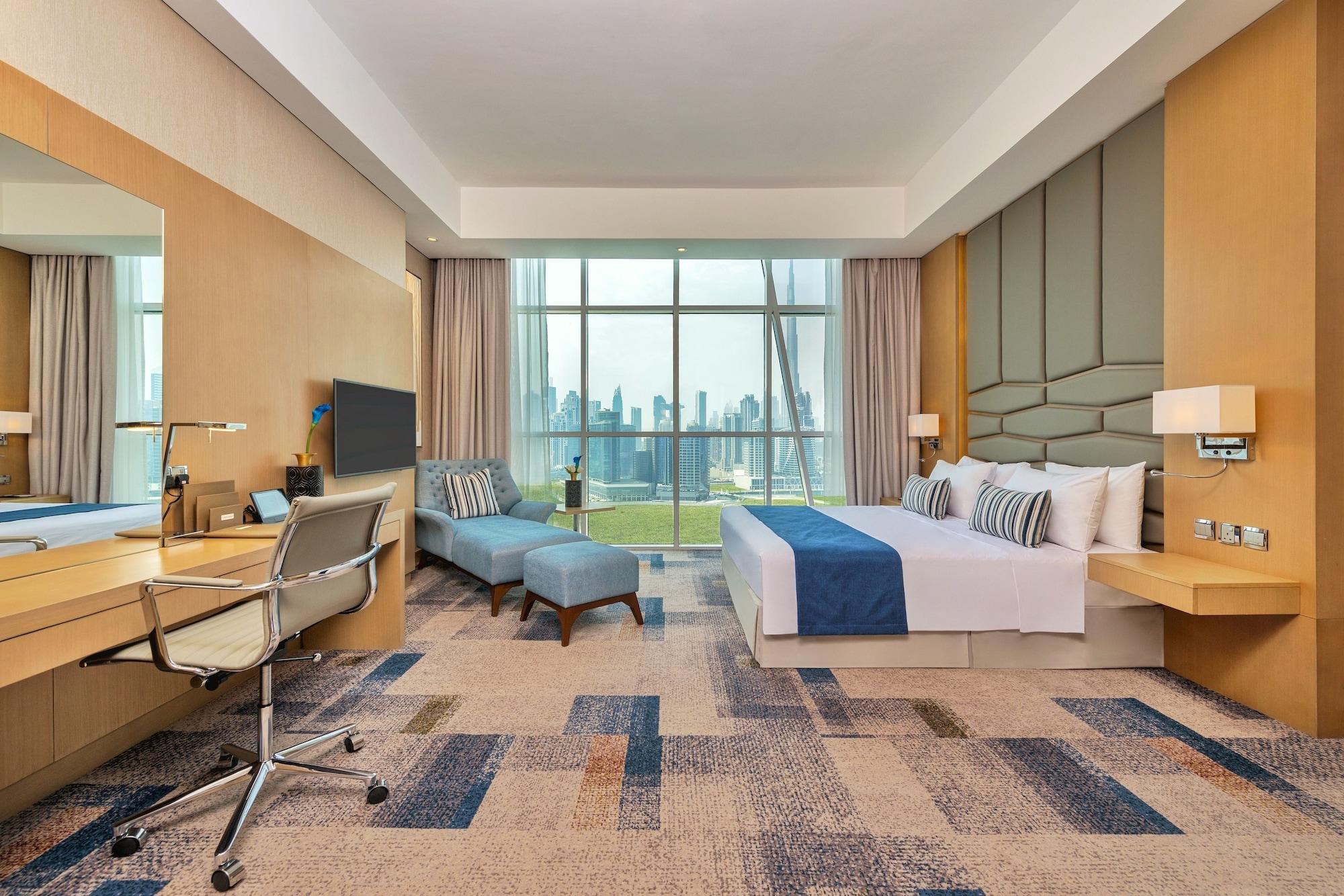 Canal Central Hotel Business Bay Dubai Exterior photo The photo shows a modern hotel room that features a large window with a view of a city skyline. The room has a light color scheme, with beige and blue accents. There is a king-sized bed with decorative pillows, a comfortable lounge chair, and a small