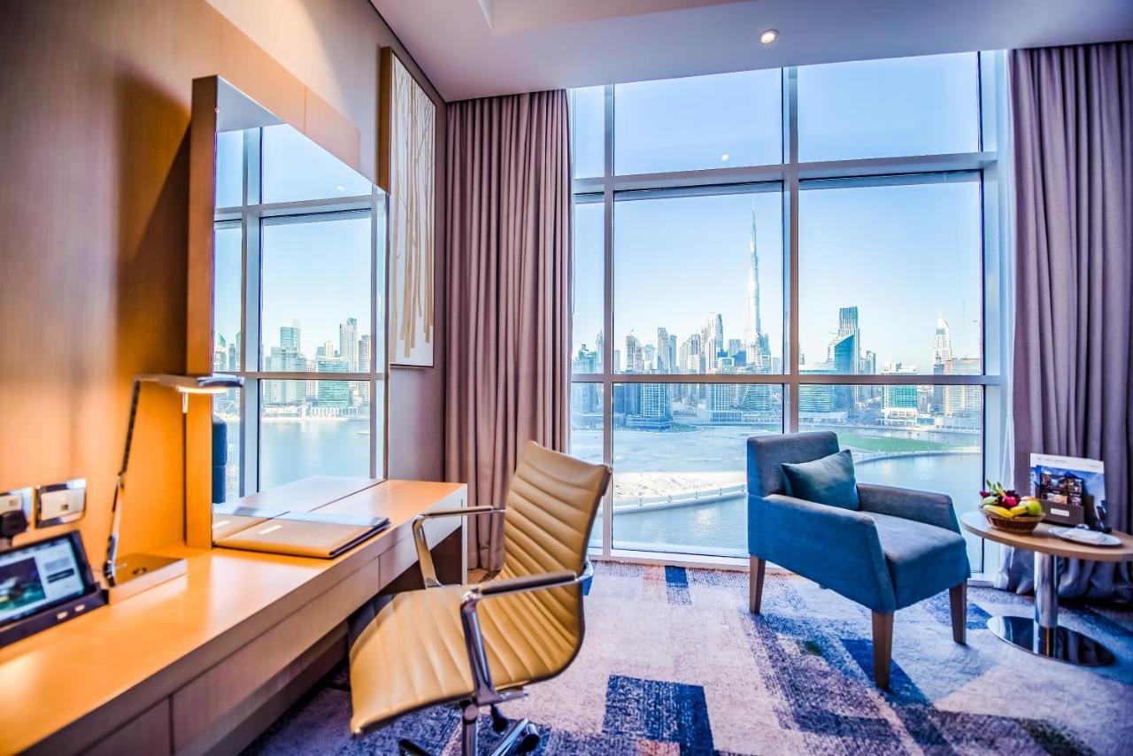 Canal Central Hotel Business Bay Dubai Exterior photo The photo depicts a modern hotel room featuring large windows that provide a panoramic view of a city skyline by the water. The interior design includes a sleek wooden desk with a comfortable, swiveling chair, and a cozy blue armchair positioned next