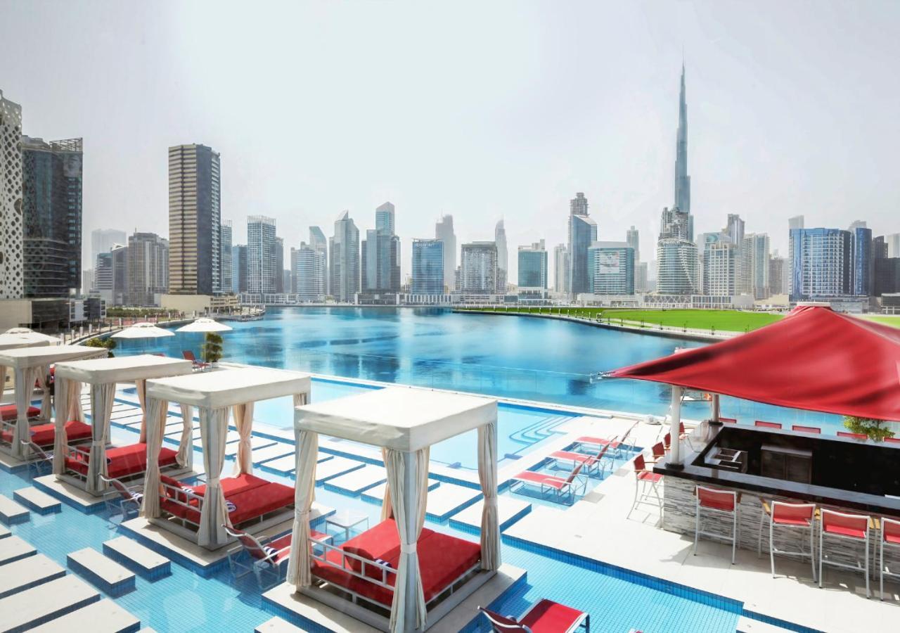 Canal Central Hotel Business Bay Dubai Exterior photo The photo shows an urban landscape featuring a pool area with white cabanas and red lounge chairs. In the background, there is a large body of water surrounded by modern skyscrapers, showcasing a city skyline. Notably, the Burj Khalifa, one of the ta