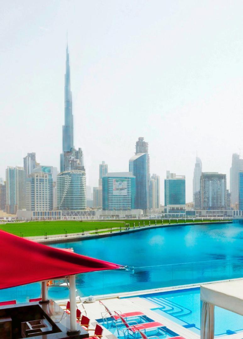 Canal Central Hotel Business Bay Dubai Exterior photo The photo showcases a modern city skyline, highlighting various skyscrapers and tall buildings, with a prominent one that appears to be the Burj Khalifa, the tallest building in the world. In the foreground, there is a body of water that reflects the