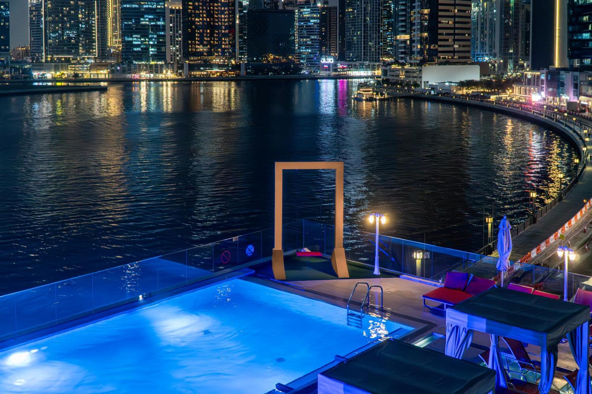 Canal Central Hotel Business Bay Dubai Exterior photo The photo shows a nighttime view of a modern pool area overlooking a body of water, likely a marina or waterfront. The pool is illuminated with a blue light, and there are lounge chairs beside it. In the background, a city skyline can be seen with ta