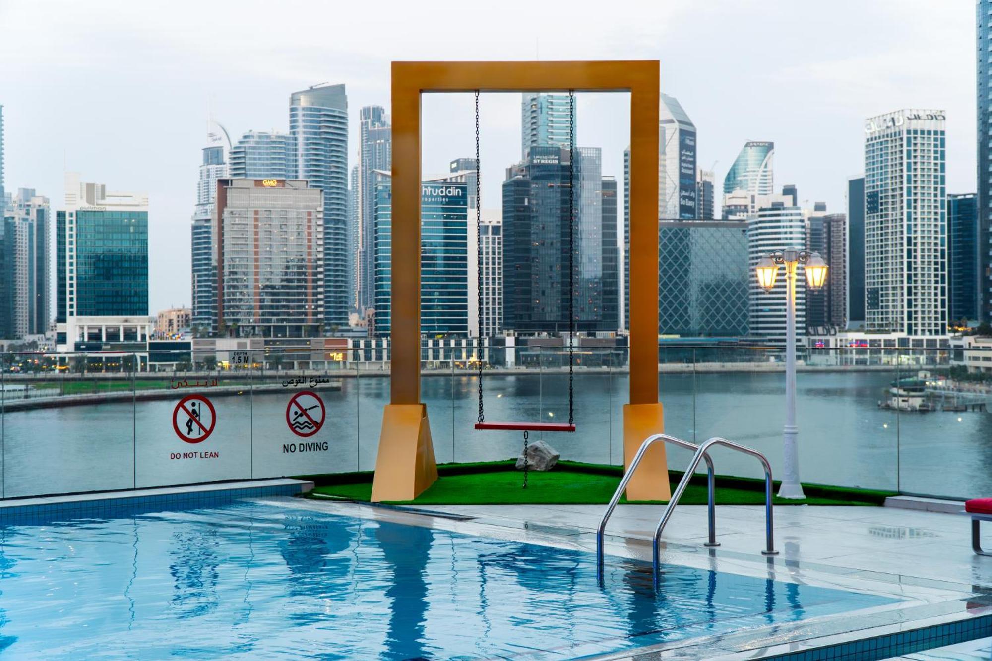 Canal Central Hotel Business Bay Dubai Exterior photo The photo depicts a modern rooftop pool area featuring a clear blue swimming pool with a ladder. In the background, there are tall city buildings, indicating a vibrant urban setting. Prominently, a large, yellow swing set is positioned next to the po