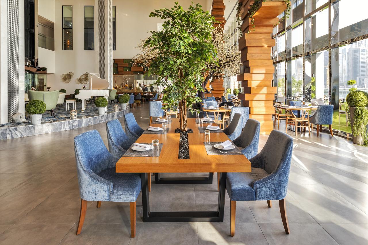 Canal Central Hotel Business Bay Dubai Exterior photo The photo shows a modern restaurant interior featuring a spacious dining area. In the center, there is a large wooden table surrounded by upholstered blue chairs. A tall, green plant is positioned in the middle of the table, adding a natural element 