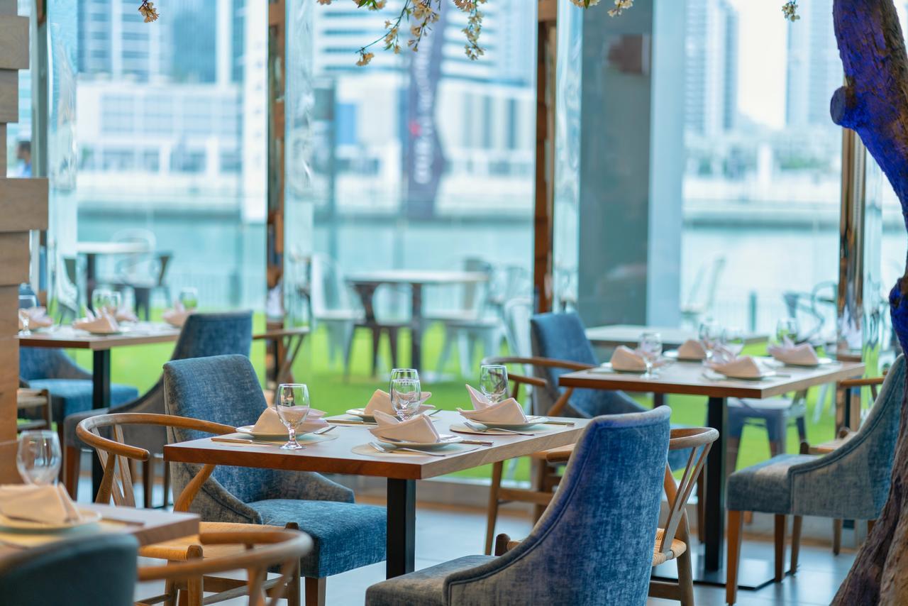 Canal Central Hotel Business Bay Dubai Exterior photo The image shows a modern restaurant interior featuring tables set with dinnerware, glasses, and napkins. The furnishings include elegant blue chairs and wooden tables. Large glass windows provide a view of the outdoors and a body of water, contributi