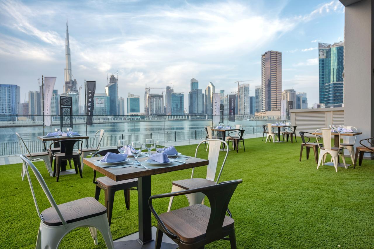 Canal Central Hotel Business Bay Dubai Exterior photo The image shows an outdoor dining area with several tables set on a grassy lawn. Each table is arranged with blue napkins and table settings, and there are a mix of modern chairs. In the background, a skyline of tall buildings, including a prominent 