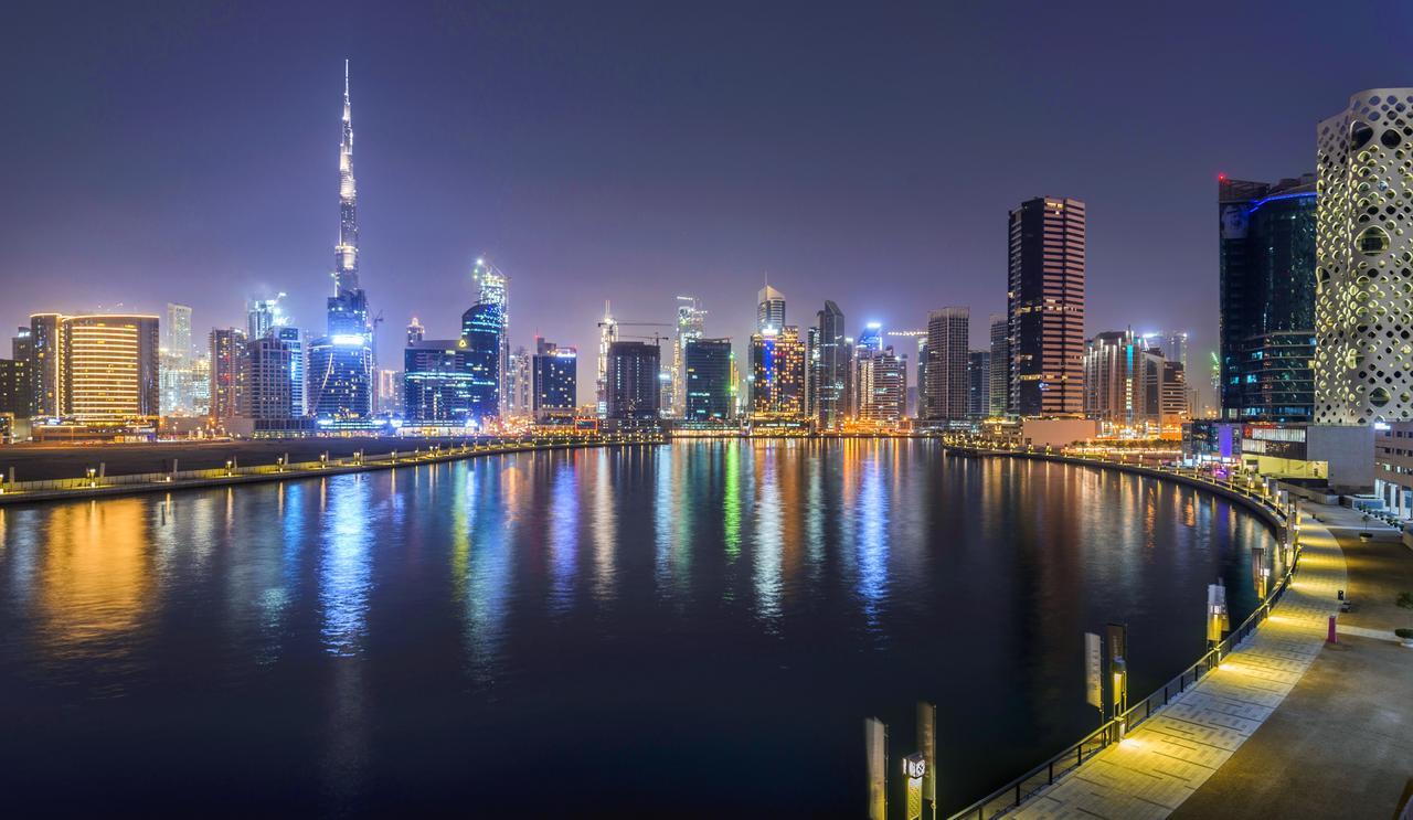 Canal Central Hotel Business Bay Dubai Exterior photo The photo shows a nighttime cityscape featuring a skyline illuminated by numerous lights. A prominent skyscraper, likely the Burj Khalifa, stands tall among other modern buildings. The scene includes a body of water reflecting the city lights, contri