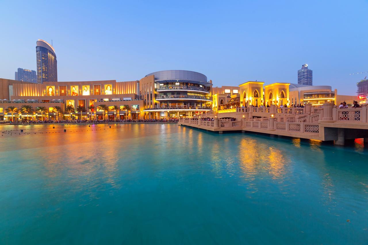 Canal Central Hotel Business Bay Dubai Exterior photo The photo depicts a scenic waterfront view, likely in an urban area during twilight. There's a large, modern building with bright lights, which seems to house shops or restaurants, alongside stylish architecture. The water in the foreground is calm a